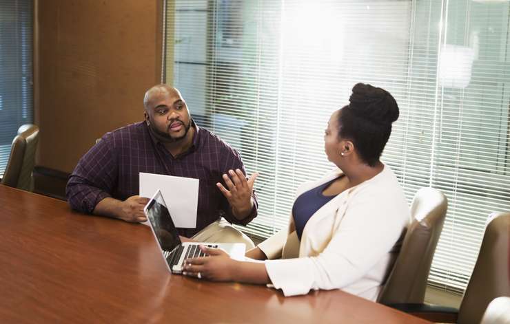 Two African-American business people having a meeting in a corporate board room. The mid adult woman in her 30s is using a laptop. The mature man in his 40s looks serious as he talks.