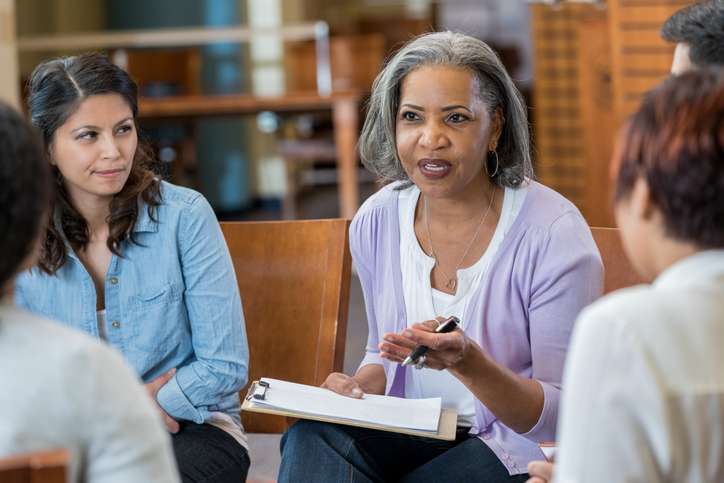 Serious senior African American female mental health profession gestures while talking with clients in a support group or group therapy session. She is holding a clipboard.