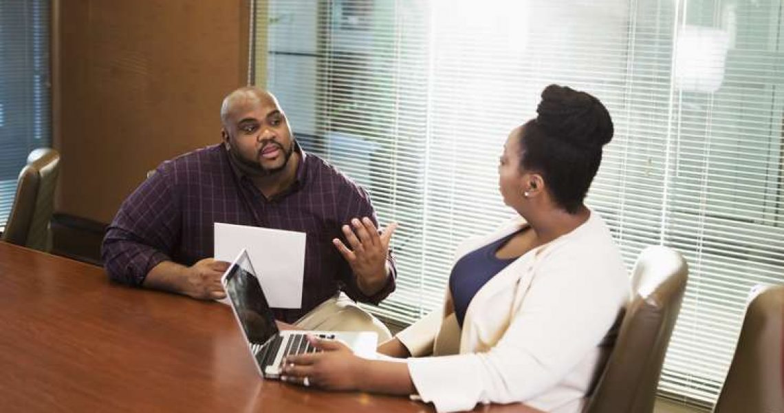 Two African-American business people having a meeting in a corporate board room. The mid adult woman in her 30s is using a laptop. The mature man in his 40s looks serious as he talks.